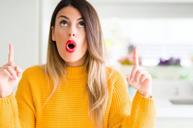 Evde kış kazak giyen genç güzel kadın hayrete ve yukarıya bakıyor ve parmakları ile işaret ve silah kaldırdı şaşırttı.