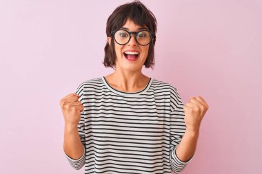 İzole pembe arka plan üzerinde çizgili t-shirt ve gözlük giyen genç güzel kadın şaşırmış ve kollarını kaldırdı ve açık gözleri ile başarı için şaşırttı. Kazanan kavramı.