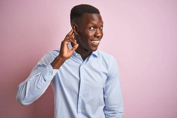 African american man wearing blue elegant shirt standing over isolated pink background smiling with hand over ear listening an hearing to rumor or gossip. Deafness concept.