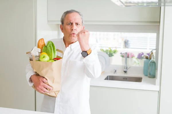 Handsome senior man holding a paper bag of fresh groceries at the kitchen annoyed and frustrated shouting with anger, crazy and yelling with raised hand, anger concept