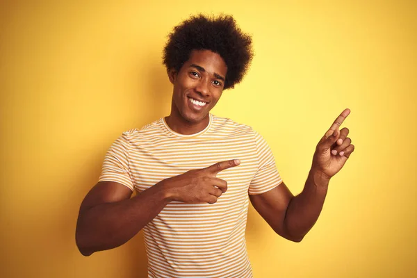 孤立した黄色の背景の上に立つストライプのTシャツを着たアメリカ人男性が微笑み 両手と指で側面を指差すカメラを見ている — ストック写真