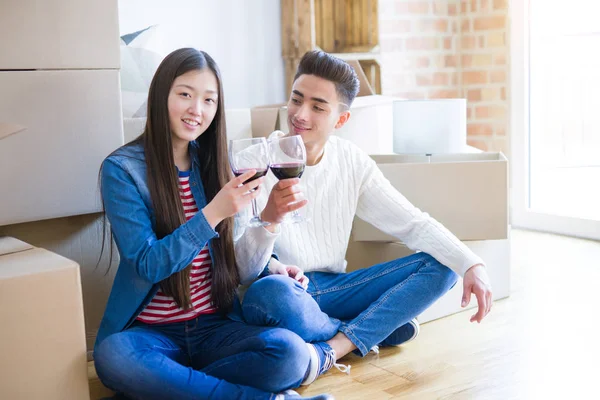 Young asian couple sitting on the floor of new apartment arround cardboard boxes, smiling drinking a glass of wine excited for moving to a new house