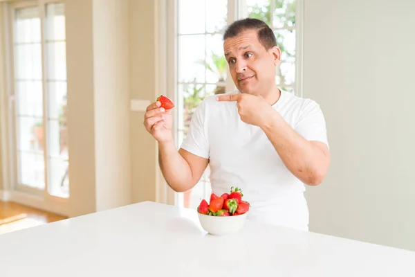 Middle age man eating strawberries at home very happy pointing with hand and finger