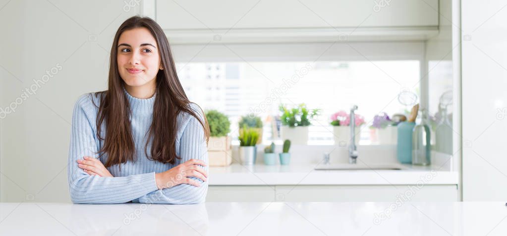 Wide angle picture of beautiful young woman sitting on white table at home smiling looking side and staring away thinking.