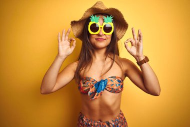 Bikini ve ananas güneş gözlüğü takan tatilde olan kadın izole sarı arka plan üzerinde rahatlayın ve parmakları ile meditasyon hareketi yaparken kapalı gözleri ile gülümseyerek. Yoga konsepti.