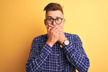 Izole sarı arka plan üzerinde rahat gömlek ve gözlük giyen genç yakışıklı adam hata için elleri ile ağız kapsayan şok. Gizli kavram.