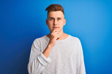 Genç yakışıklı adam beyaz rahat t-shirt giyen izole mavi arka plan ciddi yüz soru hakkında düşünme ayakta, çok karışık bir fikir