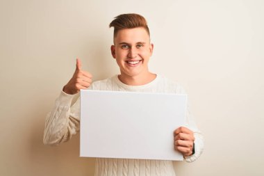 Genç yakışıklı adam büyük bir gülümseme ok işareti yapıyor mutlu izole beyaz arka plan üzerinde duran afiş tutan, parmakları ile başparmak, mükemmel işareti