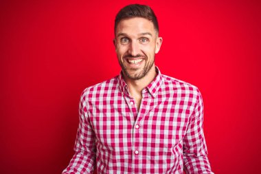 Kırmızı izole arka plan üzerinde genç yakışıklı adam samimi karşılama, olumlu ve kendinden emin selamlar gibi açık kolları ile neşeli gülümseyen