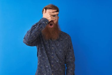 Genç kızıl saçlı İrlandalı adam izole mavi arka plan üzerinde duran çiçek yazlık gömlek giyen şok el ile yüz ve gözleri kapsayan gözetleme, utanmış ifade ile parmaklarından bakarak.