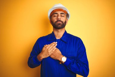 Izole sarı arka plan üzerinde üniforma ve kask giyen yakışıklı Hint işçi adam kapalı gözleri ve yüzünde minnettar jest ile göğüs üzerinde elleri ile gülümseyerek. Sağlık kavramı.