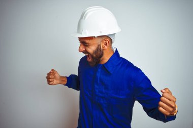 Yakışıklı hint işçi adam izole beyaz arka plan üzerinde üniforma ve kask giyen çok mutlu ve heyecanlı kolları kaldırdı ile kazanan jest yapıyor, gülümseyerek ve başarı için çığlık. Kutlama konsepti.