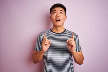 İzole pembe arka plan üzerinde duran çizgili t-shirt giyen genç Asya çinli adam şaşırttı ve yukarı bakarak ve parmakları ile işaret ve kolları kaldırdı şaşırttı.