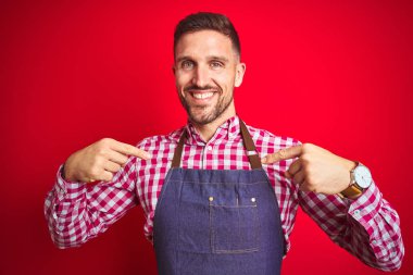 Genç yakışıklı adam kırmızı izole arka plan üzerinde dükkan sahibi önlük üniforma giyen yüzünde gülümseme ile emin görünümlü, parmakları gururlu ve mutlu kendini işaret.