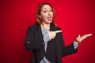 İzole kırmızı arka plan üzerinde zarif ceket giyen güzel kızıl saçlı iş kadını şaşırttı ve el ile sunarken kameraya gülümseyerek ve parmak ile işaret.