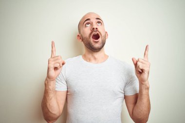 Sakallı genç kel adam izole arka plan üzerinde rahat beyaz t-shirt giyen şaşırttı ve yukarı bakarak ve parmakları ile işaret ve kolları kaldırdı şaşırttı.
