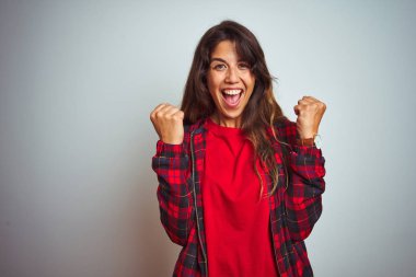 Kırmızı tişört ve ceket giyen genç güzel kadın beyaz izole arka plan üzerinde duran sürpriz ve kollarını kaldırdı ve açık gözleri ile başarı için şaşırttı. Kazanan kavramı.