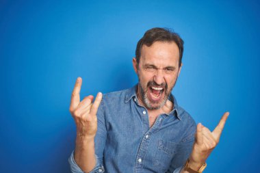 Izole mavi arka plan üzerinde gri saçlı Yakışıklı orta yaş yaşlı adam elleri yukarı ile rock sembolü yapıyor deli ifade ile bağırıyor. Müzik yıldızı. Ağır müzik konsepti.