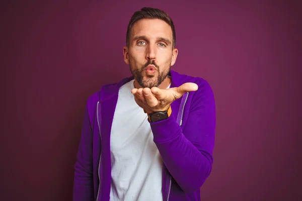 Young fitness man wearing casual sports sweatshirt over purple isolated background looking at the camera blowing a kiss with hand on air being lovely and sexy. Love expression.