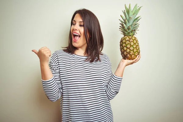 年轻的美丽女人拿着热带水果菠萝在孤立的背景指向和显示与大拇指到一边与快乐的脸微笑 — 图库照片