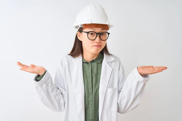 Young chinese engineer woman wearing coat helmet glasses over isolated white background clueless and confused expression with arms and hands raised. Doubt concept.