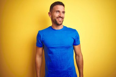 Genç yakışıklı adam sarı izole arka plan üzerinde rahat mavi t-shirt giyen yüzünde gülümseme, doğal ifade ile yan bakıyor. Gülmekten emin.