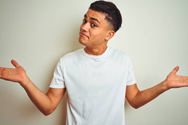 Genç Brezilyalı adam izole beyaz arka plan üzerinde duran t-shirt giyen habersiz ve kolları ve elleri ile karışık ifade kaldırdı. Şüphe kavramı.