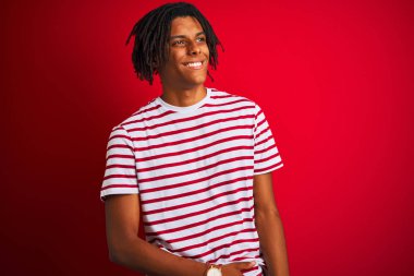 Çizgili t-shirt giyen dreadlocks giyen genç afro adam yüz, doğal ifade gülümseme ile yan bakan izole kırmızı arka plan üzerinde duruyor. Gülmekten emin.