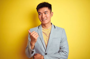 Genç Asyalı Çinli işadamı izole sarı arka plan üzerinde duran ceket giyen mutlu yüz görünümlü gülümseyerek ve başparmak ile yan işaret.