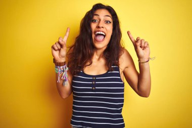 İzole sarı arka plan üzerinde duran çizgili t-shirt giyen genç güzel kadın şaşırmış ve şaşırttı ve parmakları ile işaret ve kolları kaldırdı.