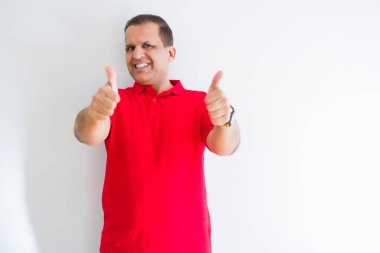 Beyaz duvar üzerinde kırmızı t-shirt giyen orta yaş adam el ile olumlu jest yapıyor onaylayan, gülümseyerek kadar başparmak ve başarı için mutlu. Kameraya bakmak, kazanan jest.