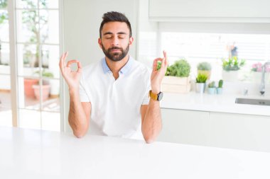 İspanyol yakışıklı casual beyaz t-shirt evde dinlenmek ve gülümseyen gözlerle parmak ile meditasyon jest yapıyor kapalı. Yoga kavramı.