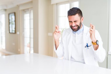 Yakışıklı İspanyol doktor ya da terapist adam klinikte tıbbi ceket giyen umut ve gözleri kapalı ile geçiş parmakları gülümseyerek. Şans ve batıl kavram.