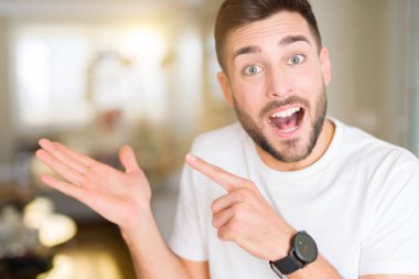 Casual beyaz t-shirt evde hayran giyen ve el ile sunulması ve parmak ile işaret ederken kameraya gülümseyen genç yakışıklı adam.