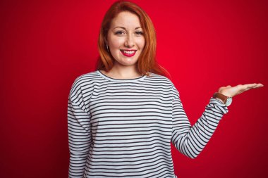 Kayışlar lacivert gömlek giyen genç kızıl saçlı kadın kırmızı izole arka plan üzerinde duran neşeli sunan gülümseyerek ve kameraya bakarak avuç içi ile işaret.