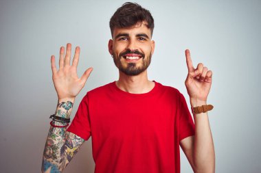 Kırmızı t-shirt giyen dövme giyen genç adam izole beyaz arka plan üzerinde duran gösteren ve parmakları ile işaret ederken kendinden emin ve mutlu gülümseyerek altı numara.