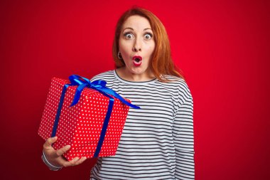 Genç güzel kızıl saçlı kadın kırmızı izole arka plan üzerinde doğum günü hediyesi tutan sürpriz bir yüz ile şok korkmuş, korku ve korku ifade ile heyecanlı