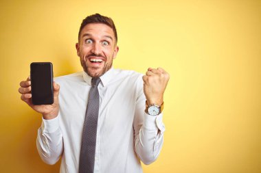 Genç yakışıklı iş adamı sarı izole arka plan üzerinde akıllı telefon ekranı gösteren gurur çığlık ve zafer ve başarı çok heyecanlı kutluyor, duygu tezahürat