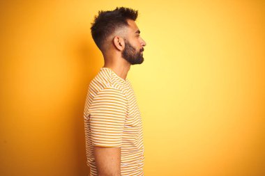 Yan bakarak izole sarı arka plan üzerinde duran t-shirt giyen genç hintli adam, doğal yüz ve kendine güvenen gülümseme ile profil poz dinlenmek.