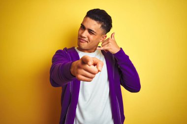 Mor kazak giyen genç brezilyalı adam izole sarı arka plan üzerinde duran telefon jesti konuşurken gülümseyerek ve sizi işaret ediyor. Beni ara.