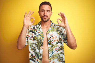Sarı izole arka plan üzerinde yaz hawaii çiçek gömlek giyen yakışıklı adam dinlenmek ve parmakları ile meditasyon jest yapıyor kapalı gözleri ile gülümseyerek. Yoga konsepti.