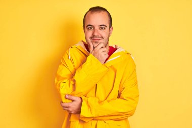 İzole sarı arka plan üzerinde duran yağmurluk giyen genç adam, kameraya güvenerek çapraz kolları ve elleri çenesi üzerinde kaldırdı. Olumlu düşünme.