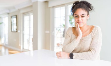 Güzel genç afro-amerikan kadın afro saçlı evde diş ağrısı veya diş hastalığı nedeniyle ağrılı ifade ile el ile ağız dokunmadan evde masada oturan. Diş hekimi konsepti.