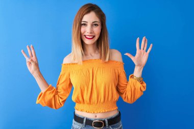 Turuncu rahat t-shirt giyen güzel redhead kadın izole mavi arka plan üzerinde duran gösteren ve parmakları ile işaret ederken emin ve mutlu gülümseyerek sekiz.