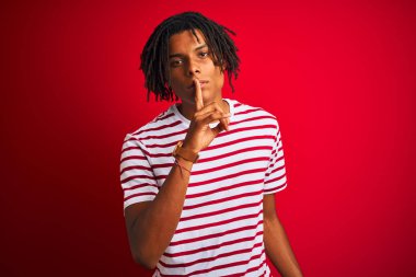 Çizgili t-shirt giyen dreadlocks ile genç afro adam dudaklarında parmak ile sessiz olmak isteyen izole kırmızı arka plan üzerinde duruyor. Sessizlik ve gizli kavram.