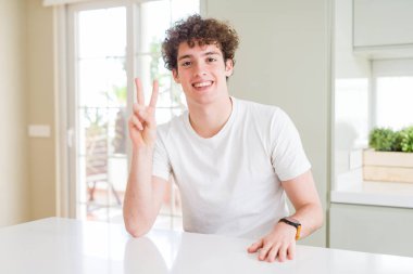 Genç yakışıklı adam evde gösteren beyaz t-shirt giyen ve emin ve mutlu gülümserken parmakları ile iki numara işaret.