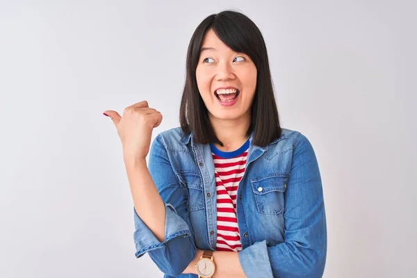 Chinese woman wearing denim shirt and red striped t-shirt over isolated white background smiling with happy face looking and pointing to the side with thumb up.