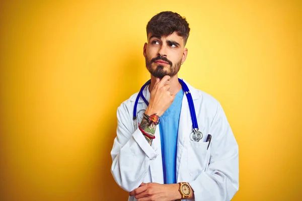 Young doctor man with tattoo wearing stethocope standing over isolated yellow background with hand on chin thinking about question, pensive expression. Smiling with thoughtful face. Doubt concept.