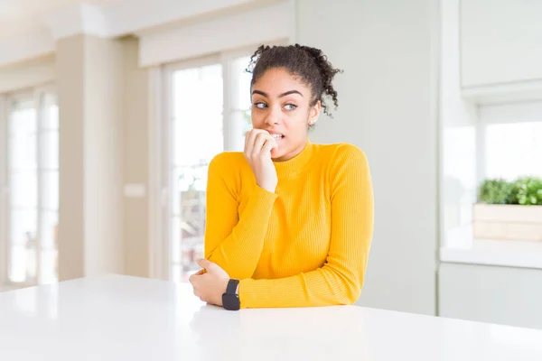 Beautiful african american woman with afro hair wearing a casual yellow sweater looking stressed and nervous with hands on mouth biting nails. Anxiety problem.