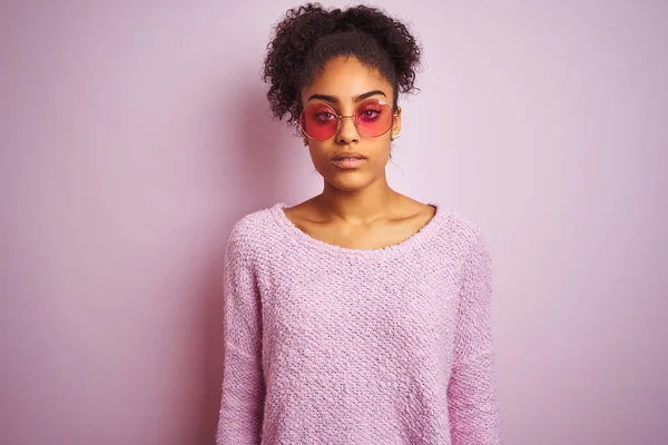 African american woman wearing winter sweater and sunglasses over isolated pink background with serious expression on face. Simple and natural looking at the camera.